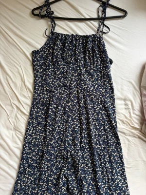 Marinblå blommig långklänning - Söt marinblå långklänning med små vita blommor och smala axelband med knyt. Klänningen har en avslappnad passform och är perfekt för varma dagar. Tillverkad i ett lätt och luftigt material som känns skönt mot huden.