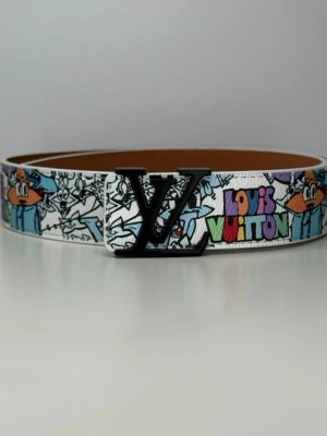  vitt bälte Looney Tunes - Exklusivt bälte i vitt skinn med färgglada Looney Tunes-figurer och stor logga i svart metall. Bältet har en unik och lekfull design med tryck i flera färger och klassisk brun insida.