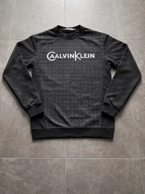 Calvin Klein sweardhirt från Calvin Klein  - Säljer en stilren och trendig Calvin Klein sweatshirt i storlek XS. Tröjan är svart med diskret CK-mönster framtill och stor vit Calvin Klein-logga över bröstet, vilket ger en exklusiv och modern look.  Den är i fint skick och passar perfekt för både vardag och en mer uppklädd streetstyle-outfit. Skön passform som sitter snyggt och bekvämt på kroppen.