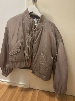 Beige bomberjacka från Neo Noir - Snygg beige bomberjacka från Neo Noir. Jackan har två snedställda fickor med lock och tryckknappar, ribbade muddar vid ärmslut och nederkant samt dragkedja framtill. Perfekt för dig som vill ha en trendig och avslappnad look.