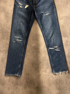 Blå slitna raka jeans med rå kant - Säljer ett par blå jeans med raka ben och flera slitningar framtill för en trendig look. Jeansen har råa, fransiga benslut och klassisk femficksmodell. Perfekta för dig som gillar en avslappnad och edgy stil.