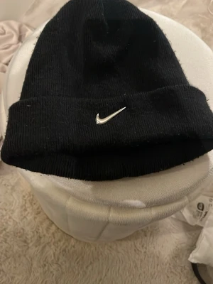 Svart Nike mössa med broderad Swoosh - Svart ribbstickad mössa från Nike med silverfärgad Swoosh-logga framtill och 'Just Do It.'-patch baktill. Klassisk uppvikt kant och mjukt material som håller dig varm. Perfekt accessoar för en sportig och stilren look.