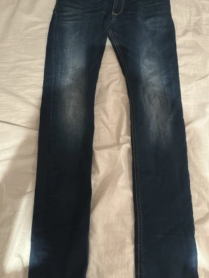 Mörkblå jeans från Diesel - Snygga mörkblå jeans från Diesel med klassisk femficksmodell och kontrastsömmar. Jeansen har en lätt tvättad look med slitningar på låren och raka ben. Perfekta för dig som gillar en tidlös och avslappnad stil. Litet hål på ena benet