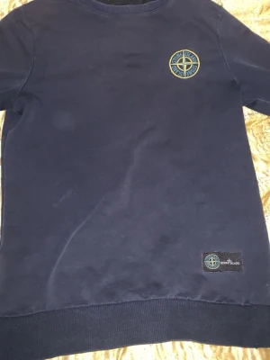 Mörkblå sweatshirt från Stone Island - Mörkblå sweatshirt från Stone Island med klassisk rund halsringning. Tröjan har det ikoniska Stone Island-märket broderat på bröstet och en patch nertill. Perfekt för en stilren och avslappnad look. Dene ganska gammal men värt att köpa.