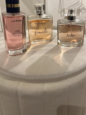 Tre parfymer: La Rive & Suddenly - Säljer tre parfymer: La Rive Her Choice med rosa vätska i en elegant rektangulär flaska med blått och guldigt lock, Suddenly Femelle i en fyrkantig flaska med guldtonad vätska och Suddenly Jolie i en liknande fyrkantig flaska med ljusrosa vätska. Alla flaskor har genomskinliga lock.