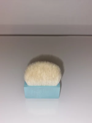 Kabukiborste med blått handtag - En kompakt kabukiborste med täta, mjuka vita borst och ett rektangulärt ljusblått handtag. Perfekt för applicering av puder eller mineralfoundation. Borsten har en modern och stilren design som gör den enkel att använda och ta med i sminkväskan.