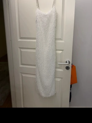 Vit långklänning med fransar - Säljer en unik vit långklänning med fransdetaljer och tunna kedjeaxelband. Klänningen har en rak passform och är perfekt för dig som vill sticka ut. Materialet känns syntetiskt och ger en cool textur. Perfekt för speciella tillfällen!