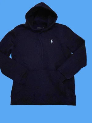 Ralph lauren hoodie - Snygg mörkblå hoodie från Polo Ralph Lauren med klassisk huva och dragsko. Ikoniska broderade loggan i vitt på bröstet och stor magficka framtill. Tillverkad i mjukt bomullsmaterial som känns skönt mot huden. Perfekt för en avslappnad och stilren look.