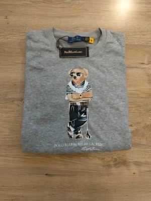 Grå sweatshirt Polo Bear Ralph Lauren - Grå sweatshirt från Ralph Lauren med ikoniskt Polo Bear-tryck framtill. Tröjan har rund halsringning, ribbade muddar och avslut. Perfekt för en avslappnad och trendig look. Det står att den är Large men den har storleken Small