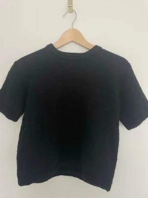 Svart stickad kortärmad topp - En svart, stickad topp med korta ärmar och rund halsringning. Modellen är croppad och har en enkel, stilren design som passar till många olika outfits. Perfekt för dig som gillar en basic men trendig look.