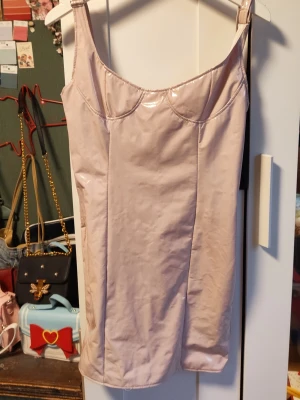 Ljusrosa kort klänning  - Säljer en ljusrosa kort klänning med smala axelband och glansig . Klänningen har en enkel, figurnära passform och sömdetaljer vid bysten. Perfekt för dig som gillar en stilren och trendig look.