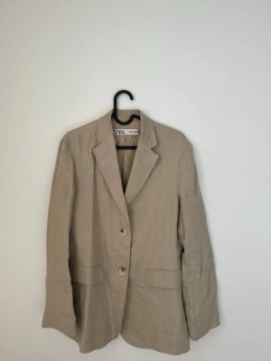 Beige kavaj från ZARA - Stilren beige kavaj från ZARA med två knappar framtill och klassisk krage. Kavajen har två fickor och en avslappnad passform, perfekt för en trendig och enkel look.