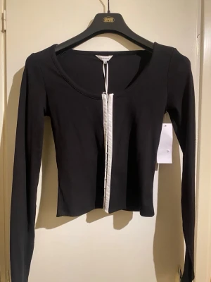 Svart ribbad croppad topp med knappar - Trendig svart croppad topp med långa ärmar och ribbad struktur. Toppen har en vit knappslå framtill som kontrast och en djup rundad ringning. Perfekt för dig som gillar stilrena detaljer och en modern look.