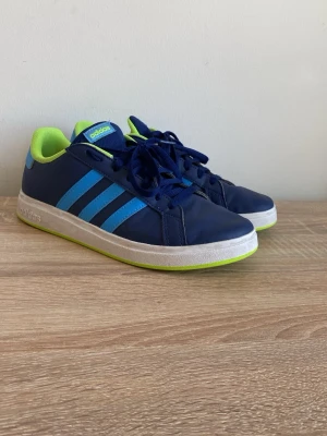 Adidas sneakers blå och lime - Snygga Adidas sneakers i mörkblått med ljusblå ränder och limegröna detaljer. Skorna har klassisk låg modell, vita sulor och ovandel i syntetmaterial. Perfekta för dig som vill sticka ut med färg och stil. Som nya men lite smuts som knappast syns på kanterna.