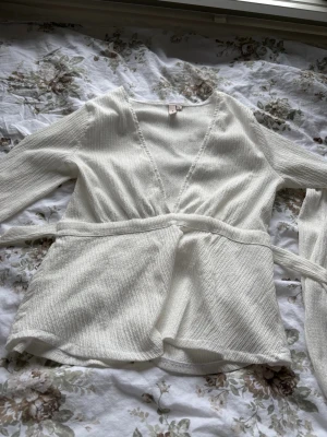 Vit urringad blus från Nelly  - Vit omlottkofta från Newbie i mjukt, strukturerat tyg. Koftan har långa ärmar, knytskärp i midjan och djup v-ringning. Enkel och stilren design som känns både chill och fräsch.