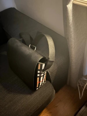 Svart axelväska från Burberry - Snygg svart axelväska från Burberry i läder med klassiskt rutigt tyg på sidorna. Väskan har ett stilrent lock och justerbar axelrem. Perfekt för dig som gillar en tidlös och trendig look med en touch av lyx.