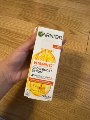 Garnier Vitamin C Glow Boost Serum - Glow Boost Serum från Garnier med vitamin C, niacinamid och salicylsyra. Förpackningen är vit och orange med en bild på en guldfärgad serumflaska. Vegansk formula som ger huden lyster och jämnare ton.
