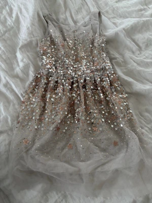 Glittrig kortklänning med paljetter - Superfin kortklänning i ljus beige med massor av glittrande silver- och guldpaljetter. Klänningen är ärmlös och har en dragkedja i ryggen. Perfekt för dig som vill sticka ut med en glittrig look. Tunt tyllmaterial ger en lätt och luftig känsla. Man kan sätta i ett band🎀 