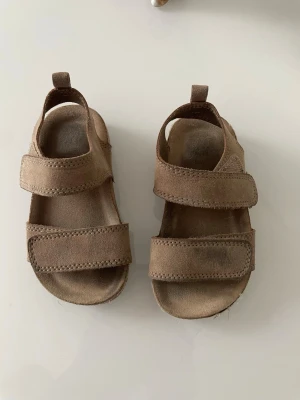 Bruna sandaler med kardborre från H&M - Snygga bruna sandaler från H&M i mockaimitation med två breda kardborreband för enkel på- och avtagning. Mjuka sulor och öppen tådesign gör dem perfekta för varma dagar. Passar till både shorts och jeans.storlek 20/21