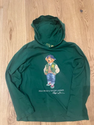 Grön hoodie Polo Bear Ralph Lauren - Mörkgrön hoodie från Ralph Lauren med Polo Bear-tryck på bröstet. Hoodien har lång ärm, huva och är gjord i mjukt bomullsmaterial. Trycket visar en nalle i collegekläder och texten 'POLO BEAR by RALPH LAUREN' under motivet.