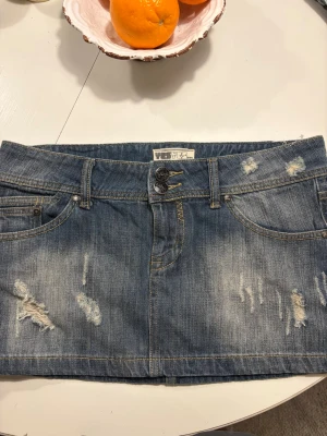 Lågmidjad Blå jeanskjol från - Säljer en kort blå jeanskjol med slitna detaljer och dubbla knappar framtill. Kjolen har klassiska fickor både fram och bak samt bälteshällor. Midjemåttet är 42cm rakt över, så ca storlek S/M💓