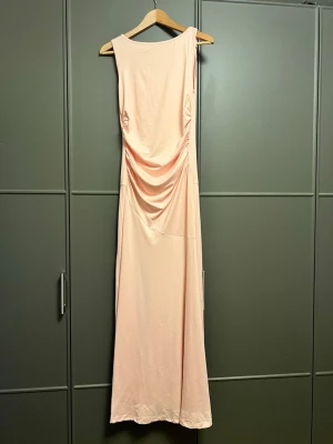 Ljusrosa långklänning med drapering - Elegant ljusrosa långklänning med snygg drapering på sidan. Klänningen har ärmlös design och rund halsringning, vilket ger en stilren och modern look. Perfekt för dig som vill ha en enkel men ändå chic klänning. Materialet känns mjukt och följsamt.