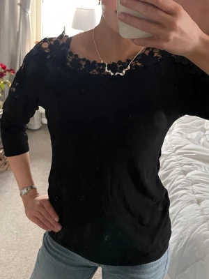 Svart topp med spetsdetaljer - Säljer en svart topp med snygga spetsdetaljer vid axlar och halsringning. Toppen har kortare ärmar. Jättebra skick i storlek 38