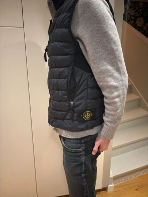 Mörkblå dunväst från Stone Island - Snygg mörkblå dunväst från Stone Island med quiltad design och dragkedja framtill. Västen har två sidofickor med dragkedja och den klassiska Stone Island-loggan på sidan. Perfekt att slänga över en hoodie eller tröja för extra värme.