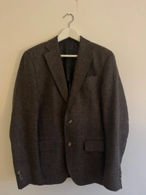 Brun kavaj i linne och ull - Snygg brun kavaj från Sand, tillverkad i en exklusiv mix av linne och ull. Kavajen har klassisk passform, två knappar framtill, fickor med lock och prickigt innerfoder. Perfekt för dig som vill ha en stilren och tidlös look.