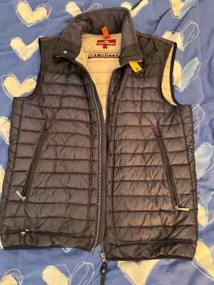 Svart dunväst från Parajumpers - Snygg svart dunväst från Parajumpers med quiltad design och två dragkedjeförsedda fickor framtill. Västen har hög krage med gul detalj och insida i beige. Perfekt för lager-på-lager och håller dig varm med lätt dunfyllning. Dragkedjan är krånglig och ett hål längst ner 