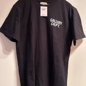Svart t-shirt från Gallery Dept. - Svart t-shirt från Gallery Dept. med vit logotyp på bröstet och stor text på ryggen. Klassisk rund hals och korta ärmar. Tillverkad i mjuk bomull för en skön känsla och enkel stil.