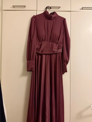 Lång vinröd klänning med knappar - Elegant långklänning i vinröd ton med hög krage och långa puffärmar. Klänningen har markerad midja, breda manschetter med dekorativa knappar och ett mjukt, flytande tyg som ger ett exklusivt intryck. Perfekt för dig som gillar klassisk och stilren design.