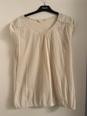 Beige topp med paljettdetaljer - Snygg beige topp med paljetter på axlarna som ger extra glans. Modellen är ärmlös och har en rund halsringning. Perfekt att styla med jeans eller kjol för en trendig look.