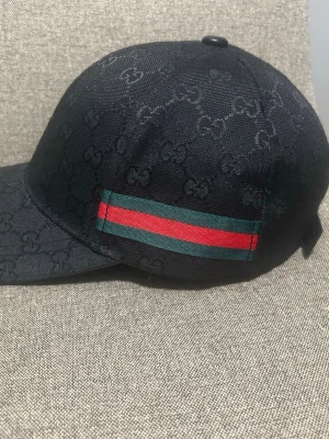 Svart keps från Gucci med GG-mönster - Snygg svart keps från Gucci med klassiskt GG-monogram över hela kepsen. På sidan finns en ikonisk grön och röd rand. Kepsen har böjd skärm och justerbar passform. Perfekt accessoar för en trendig streetwear-look.