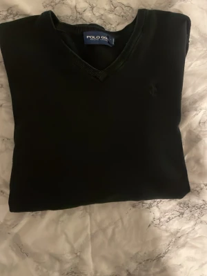 Svart V-ringad tröja Polo Golf - Klassisk svart/ mörkblå V-ringad tröja från Polo Golf Ralph Lauren. Tröjan har diskret broderad logga på bröstet och ribbade muddar vid halsringning, ärmslut och nederkant. Perfekt för en stilren look. storlek M