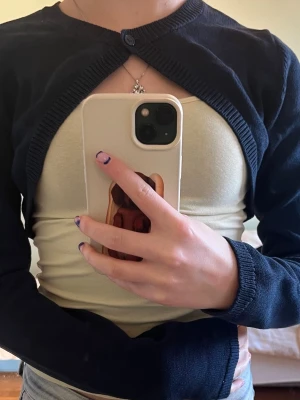 Mörkblå bolero från Gina Tricot - Kort, mörkblå bolero från Gina Tricot med lång ärm och rund halsringning. Stängs med en knapp framtill och har ribbade kanter. Perfekt att slänga över ett linne eller klänning för en snygg lager-på-lager-look.