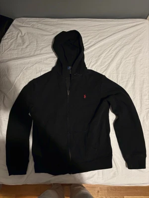 Svart hoodie från Polo Ralph Lauren - Svart hoodie med dragkedja från Polo Ralph Lauren med ett rött märke. Den är lite använd men har inga skador. Nypris: 1095kr. Passar om man är ca 170-175 cm lång. 