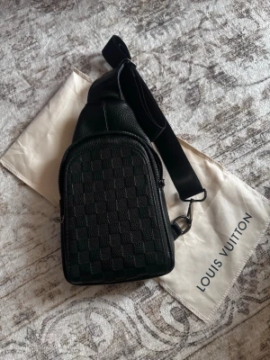 Louis Vuitton svart crossbody-väska - Snygg svart crossbody-väska från Louis Vuitton med klassiskt rutmönster sopm ger en lyxig vibe. Väskan har justerbar axelrem i textil, silverfärgade detaljer och är tillverkad i läder. Perfekt för dig som vill ha en stilren och praktisk accessoar.