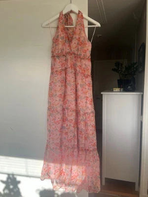 Blommig långklänning med halterneck - Säljer en romantisk långklänning med blommigt mönster i rosa, orange och ljusa toner. Klänningen har halterneck och är ärmlös, med volangdetaljer och luftig passform. Perfekt för varma dagar och sommarens alla tillfällen.