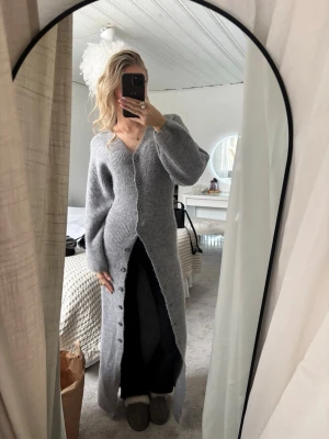 Lång grå stickad kofta  - Lång, mjuk och stickad grå cardigan med v-ringning och stora knappar framtill. Modellen har långa ärmar med lätt puff och ribbstickade detaljer. Perfekt för lager-på-lager och chill dagar.