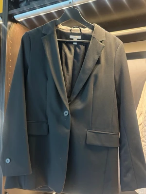 Svart klassisk kavaj med knappar från H&M - Stilren svart kavaj med två knappar framtill och klassisk krage. Kavajen har två fickor med lock och är helfodrad. Perfekt för en snygg och tidlös look. Passar till många olika outfits och har en rak, något längre passform.