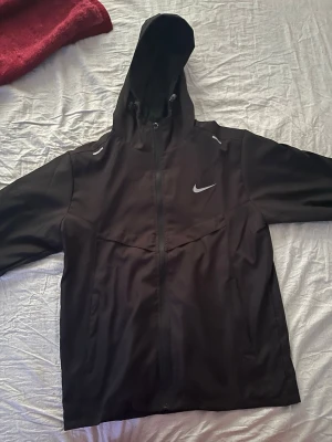 Svart Nike vindjacka med huva - Snygg svart vindjacka från Nike med hel dragkedja och justerbar huva. Jackan har reflekterande detaljer och den klassiska Nike-loggan på bröstet. Tillverkad i lätt och vindtätt material, perfekt för blåsiga dagar. Sportig och stilren design. Det är en Medium men dock är den liten i storleken så skulle rekommenderas till de som har Small