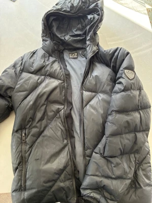 Svart pufferjacka från EA7 Armani - Snygg svart pufferjacka från EA7 Emporio Armani med huva och dragkedja framtill. Jackan har två fickor med dragkedja och ett stilrent EA7-märke på ärmen. Perfekt för kalla dagar och ger en modern look.