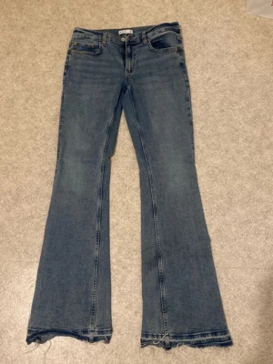 Blå bootcut jeans med rå kant - Ett par blå bootcut jeans med rå, fransig kant längst ner. Klassisk femficksmodell och normalhög midja. Jeansen har en avslappnad passform över låren och utsvängda ben. Tillverkade i denim, perfekt för en trendig och avslappnad stil.