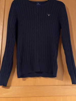 Mörkblå kabelstickad tröja Gant - Snygg mörkblå kabelstickad tröja från Gant i stretchig bomull. Tröjan har rund halsringning, diskret logga på bröstet och ribbade muddar vid ärmslut och nederkant. Perfekt för en klassisk och stilren look.