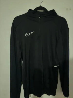 Nike academy - Svart långärmad träningströja från Nike med vit logga på bröstet och vita detaljer vid sidorna. Tröjan har halv dragkedja framtill och är tillverkad i ett lätt och funktionellt material som passar perfekt för sport och träning.