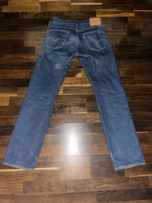 Levi's 501 - Klassiska blå jeans från Levi's med rak passform och fem fickor. Jeansen har en mörk tvätt med lätt slitning och detaljer i kopparfärgade nitar. Tillverkade i bomull med Levi's ikoniska läderpatch bak i midjan.