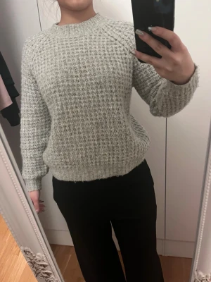 Ljusgrå stickad tröja med struktur - Mysig ljusgråt stickad tröja med rund hals och lång ärm. Tröjan har en grovstickad struktur och ribbade muddar vid ärmslut och nederkant. Perfekt för kalla dagar och enkel att matcha till olika outfits.