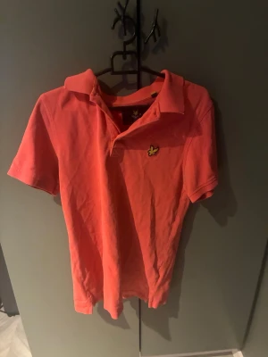 Korallröd pikétröja från Lyle & Scott - Säljer en korallröd pikétröja från Lyle & Scott med klassisk krage och korta ärmar. Tröjan har den ikoniska gula loggan broderad på bröstet och är tillverkad i bomull. Perfekt för en avslappnad och stilren look.