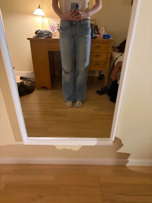 Ljusblå raka jeans med hål på knät - Säljer ett par ljusblå jeans med raka ben och ett snyggt hål på ena knät. Jeansen har klassisk femficksdesign och hög midja. Perfekta för en avslappnad och trendig stil.